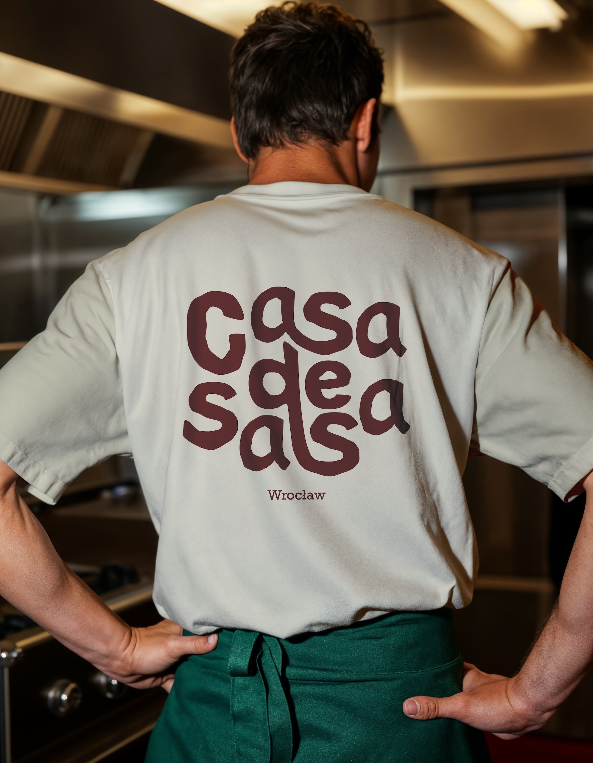 Kucharz w kuchni restauracyjnej tyłem do obiektywu, ubrany w białą koszulkę z napisem 'casa de salsa Wrocław' i zielony fartuch