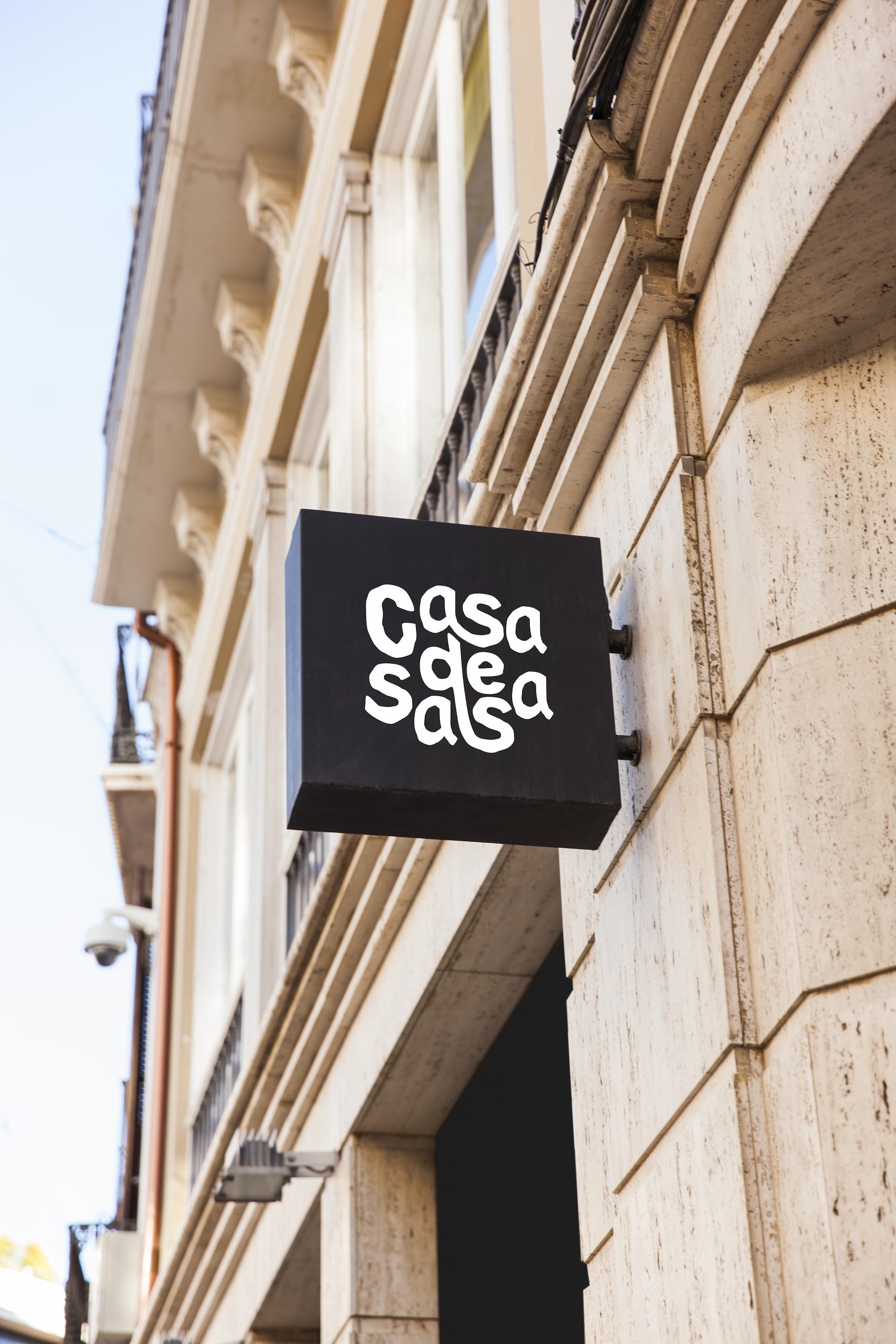 Czarny szyld z białym logo Casa de Salsa zamontowany na ścianie kamienicy – widok od dołu na tle błękitnego nieba
