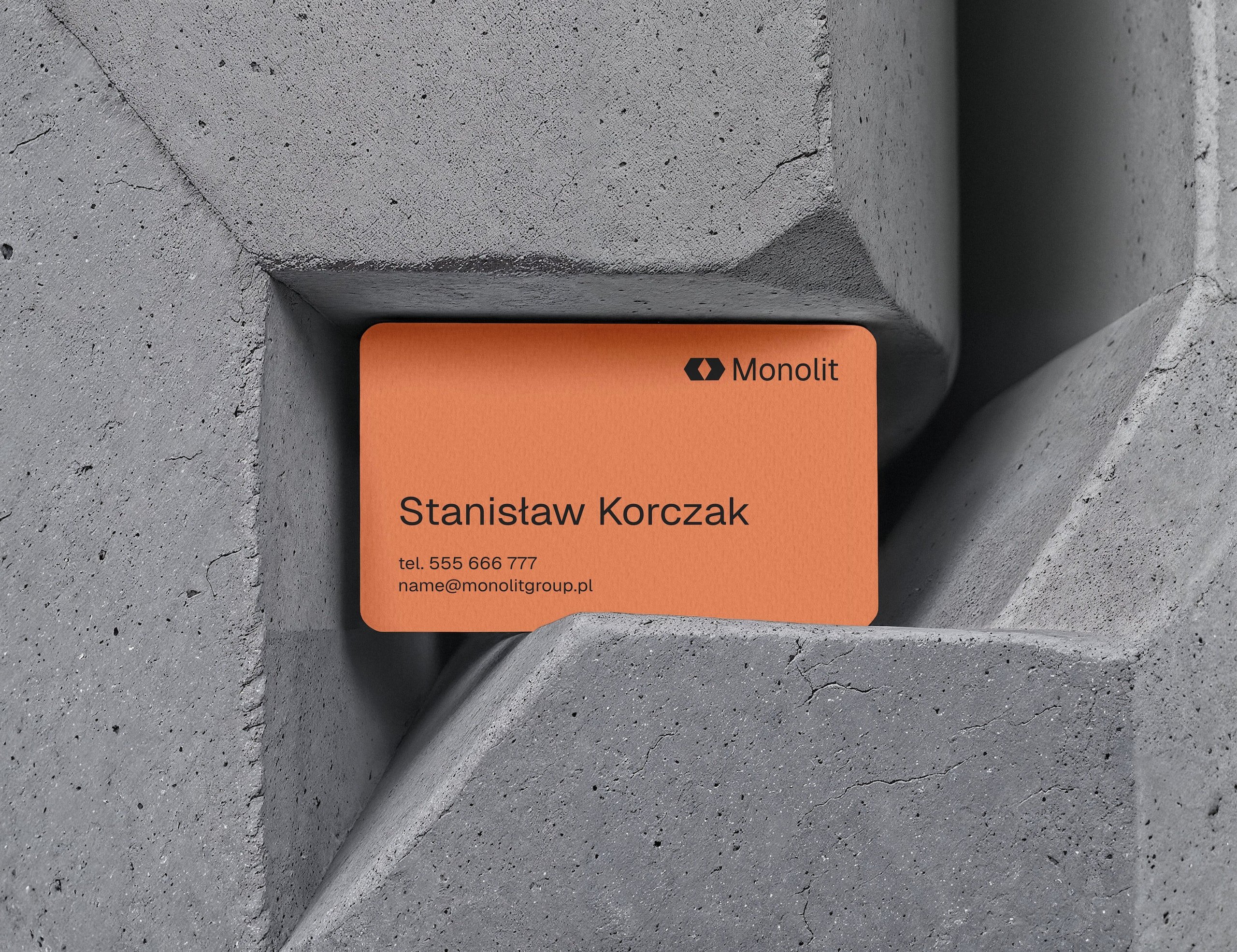 Pomarańczowa wizytówka Monolit z imieniem Stanisław Korczak wciśnięta między elementy szarej betonowej konstrukcji