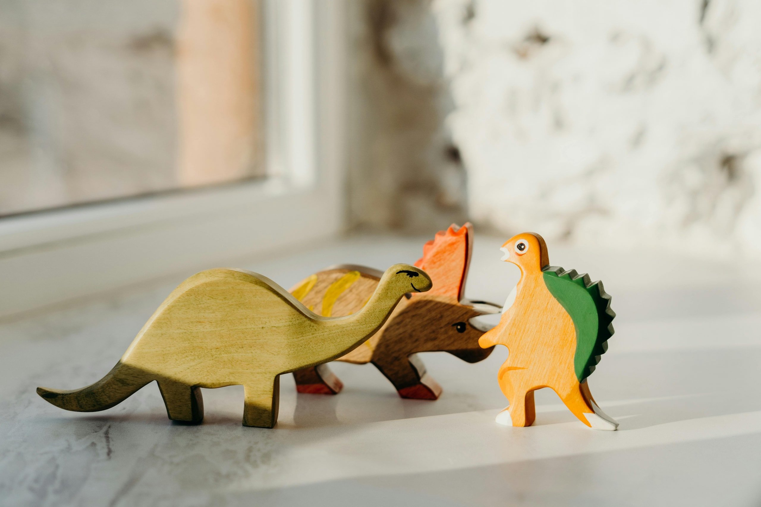 Trzy drewniane figurki dinozaurów ustawione na parapecie w promieniach słońca – brontosaurus, triceratops i spinosaurus w naturalnych barwach drewna